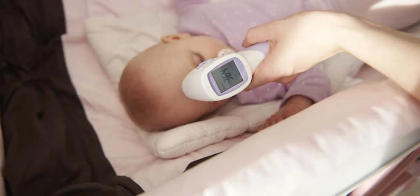 Bébé couché dans son berceau pendant qu’un adulte prend sa température avec un thermomètre infrarouge frontal.
