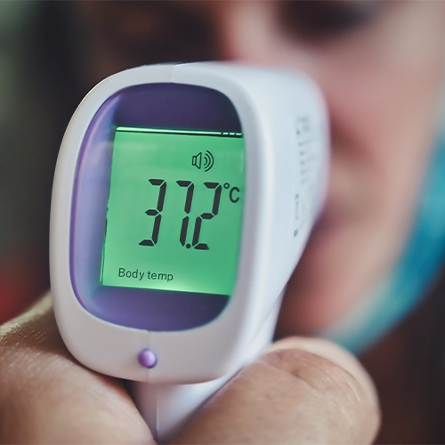 À Propos de 1 L'image montre un gros plan d'un thermomètre infrarouge sans contact blanc et violet. Son écran rétroéclairé vert affiche "37.2°C" et "Body temp" (Température corporelle), indiquant une lecture de la température corporelle. Il y a aussi une icône ressemblant à un haut-parleur avec des ondes sonores, suggérant que la fonction sonore est activée. Une main, probablement celle de la personne qui prend la température, tient l'appareil. À l'arrière-plan flou, une partie d'un visage est visible.