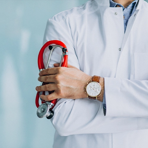 À Propos de 2 Un médecin portant une blouse blanche et une chemise bleue claire est représenté les bras croisés. Il porte une montre élégante avec un bracelet en cuir marron et un cadran blanc. Dans ses mains croisées, il tient un stéthoscope rouge et argenté. L'arrière-plan est uni et de couleur claire.