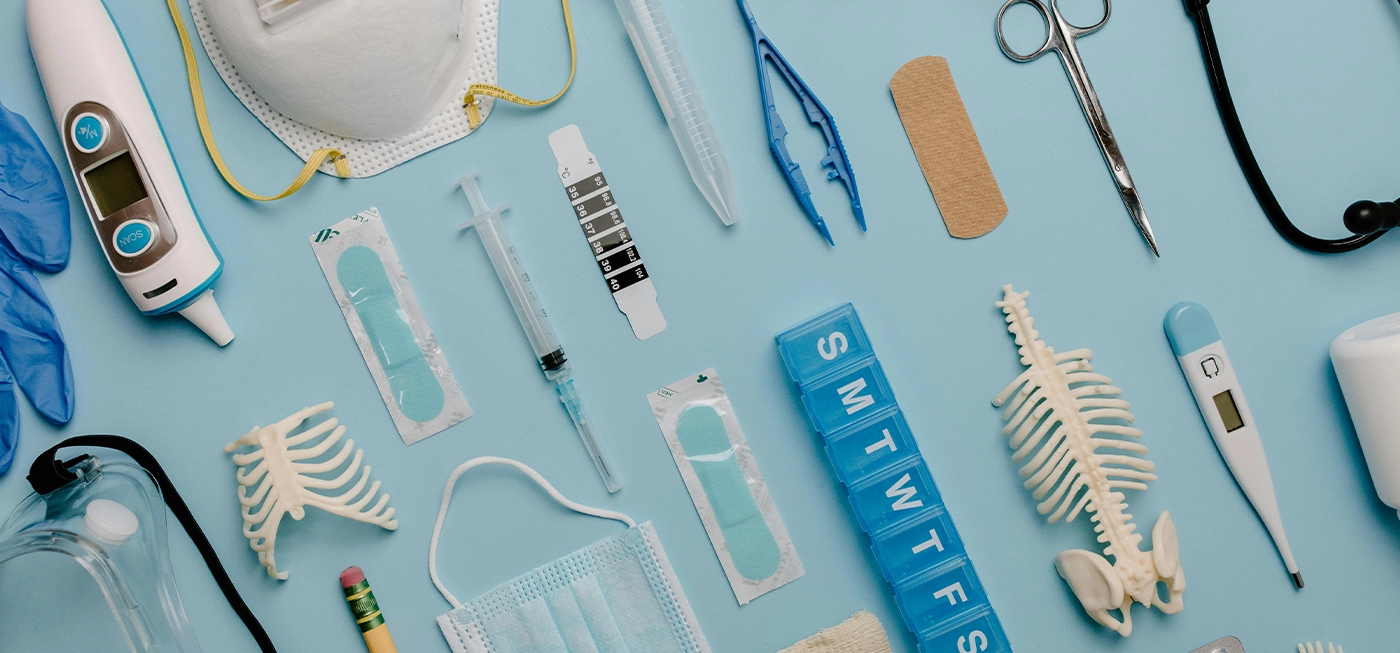 Matériel Médical en Ligne : Produits de Santé, Accessoires et Équipements de Qualité à Prix Pas Cher 4 Matériel Médical - Divers équipements médicaux disposés sur fond bleu : thermomètre frontal, seringues, masque, gants, stéthoscope, ciseaux, pilulier hebdomadaire, pansements et petits modèles de squelette.
