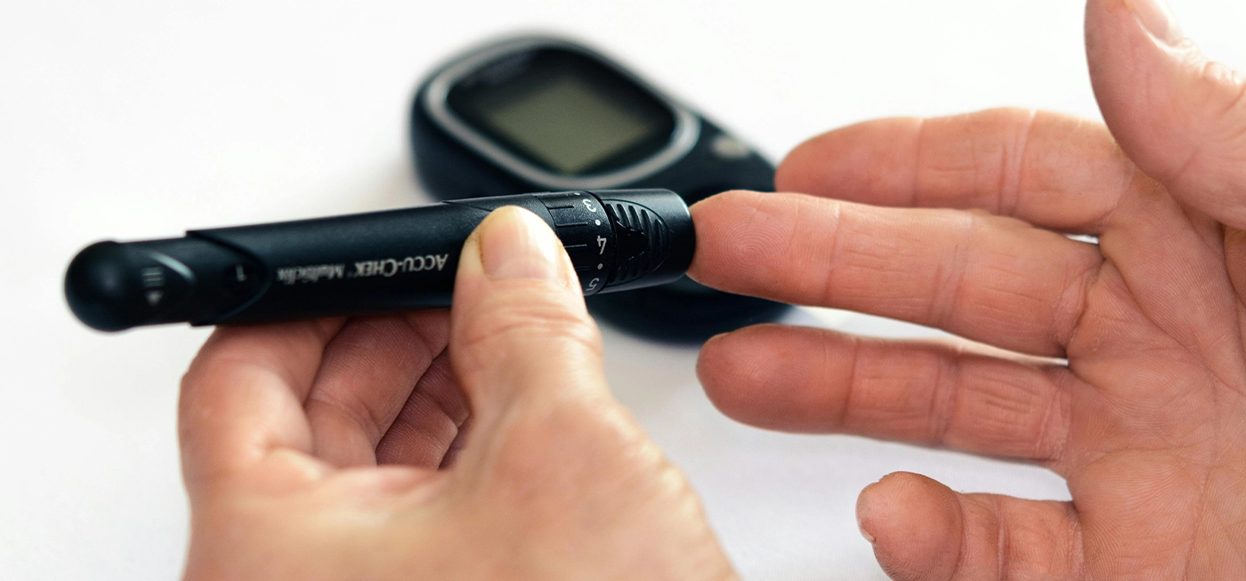 Matériel Médical en Ligne : Produits de Santé, Accessoires et Équipements de Qualité à Prix Pas Cher 3 Main d’une personne utilisant un stylo autopiqueur Accu-Chek pour tester la glycémie avec un lecteur de glycémie posé à l’arrière-plan.